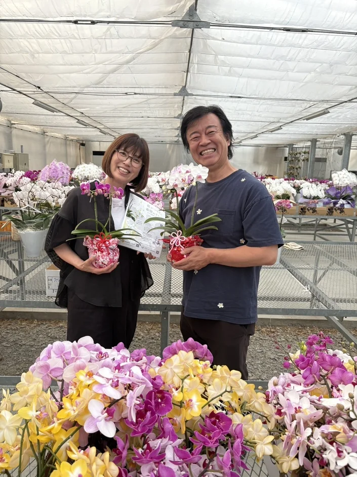 温室で満面の笑みを浮かべる男女が、それぞれ小さな胡蝶蘭の鉢植えを持っている様子です。背景には色とりどりの胡蝶蘭が並び、活気に満ちた空間が広がっています。
