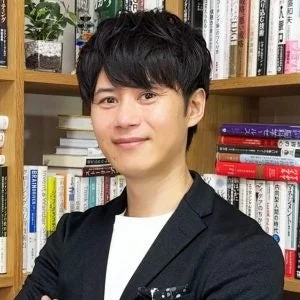 書棚を背景に、黒いジャケットと白いシャツを着た男性が微笑んでいるポートレートです。知的で親しみやすい印象を与えます。