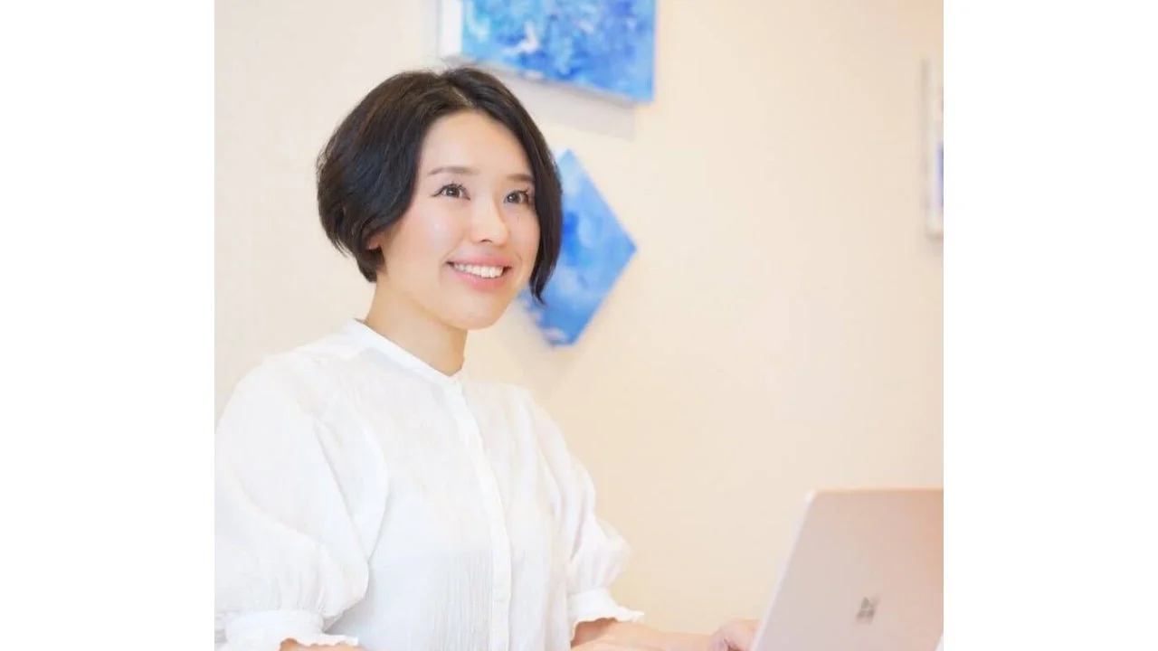 白いブラウスを着た笑顔の日本人女性がノートパソコンに向かっている画像です。背景には抽象的な青いアートが飾られており、明るく爽やかな印象を与えます。