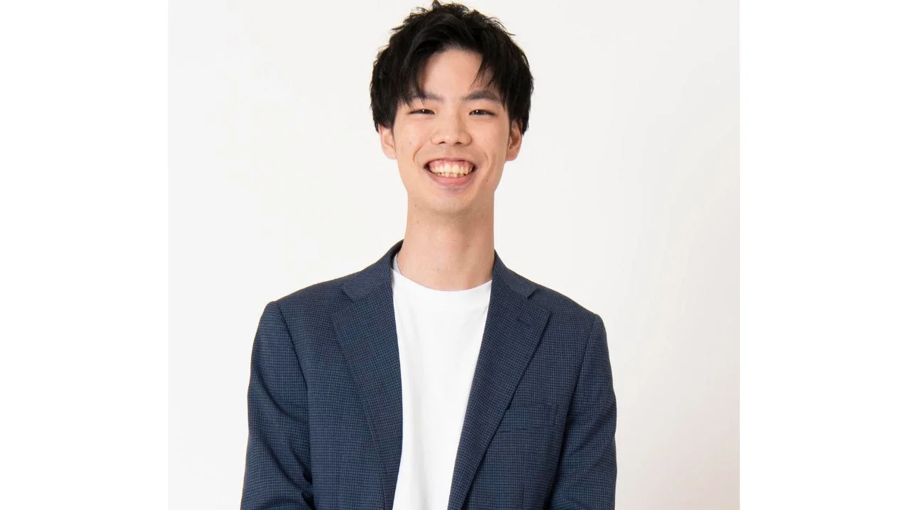 白い背景の前で、紺色のジャケットと白いTシャツを着用した若い男性が、満面の笑みを浮かべている上半身のポートレート写真です。清潔感のある明るい印象を与えます。