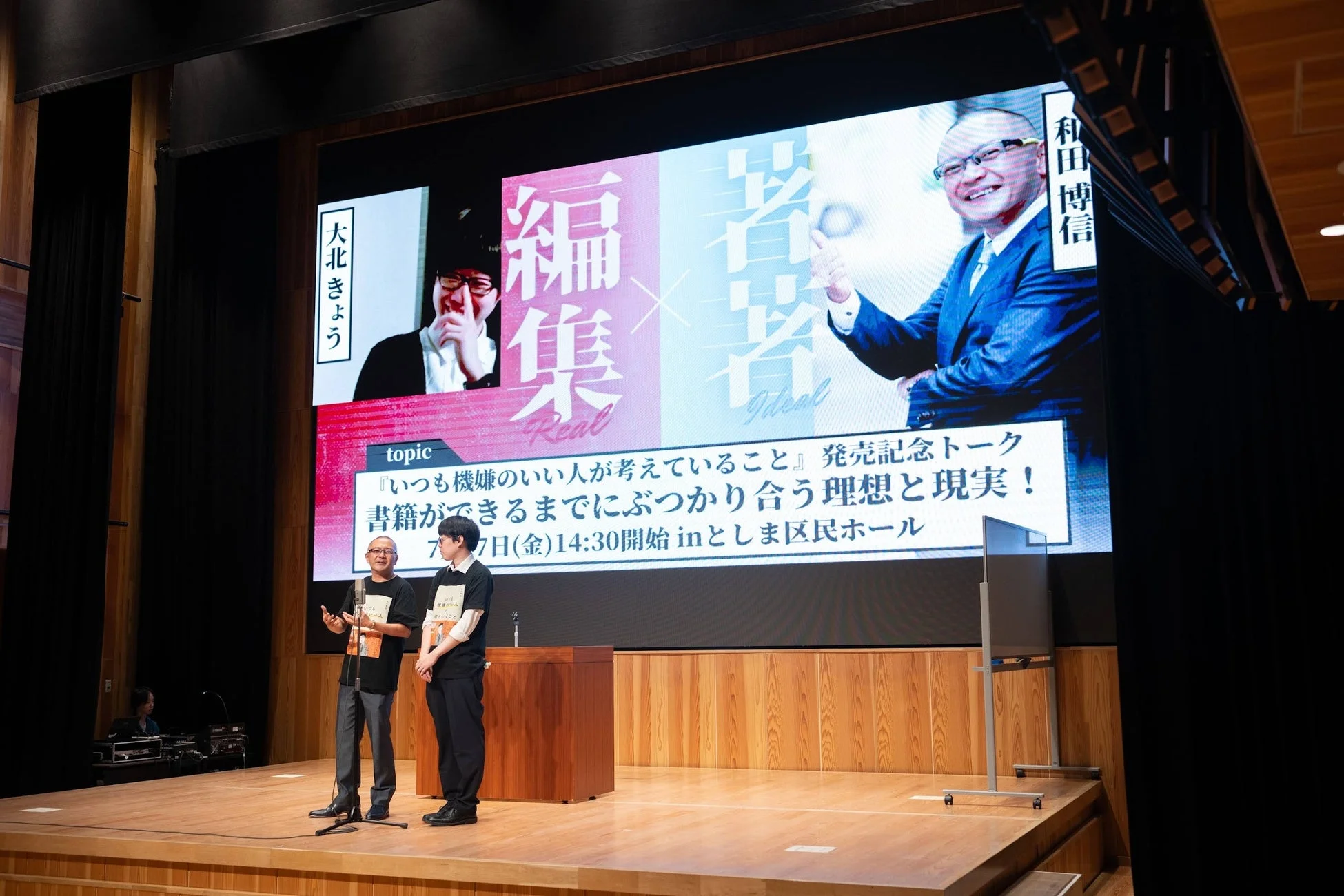 大きなスクリーンに「編集 Real」と「著者 Ideal」の文字と人物写真が映し出されたステージで、2人の男性がマイクを使ってトークをしているイベントの様子。書籍の発売記念トークショーのようだ。