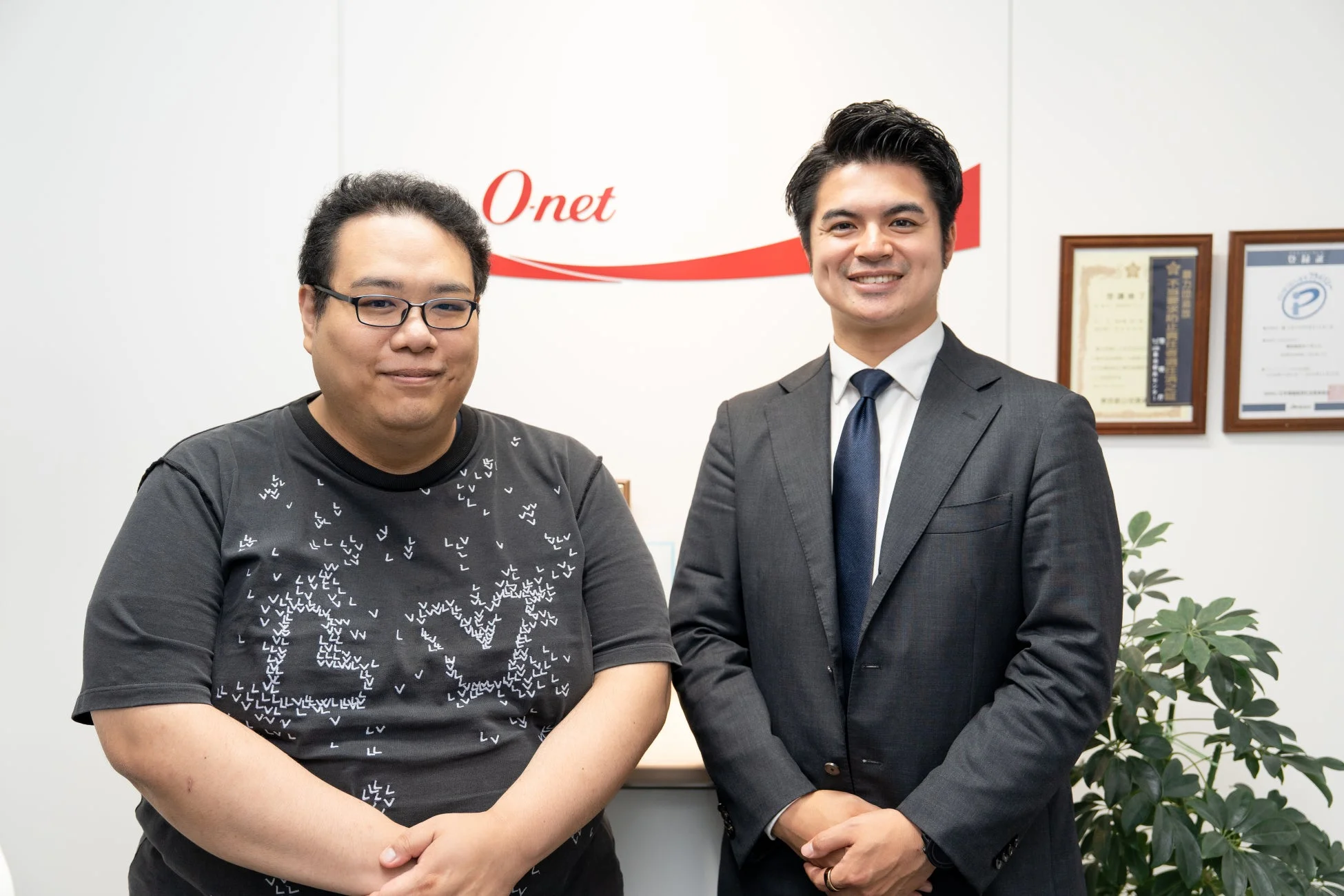 Onetのロゴがあるオフィスで、スーツ姿の男性とカジュアルな服装の男性が笑顔で並んでいます。ビジネスパートナーシップや対談の様子を捉えた写真です。