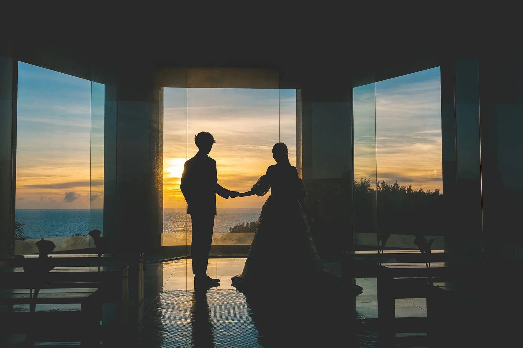 夕焼けに染まる海を背景に、大きな窓のあるチャペルで手を取り合う新郎新婦のシルエット