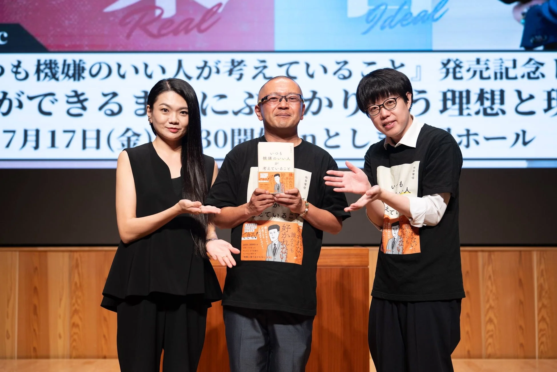 著者を含む3人がステージに立っており、書籍「いつも機嫌のいい人が考えていること」の発売記念イベントの様子です。中央の男性が本を掲げ、背景にはイベントタイトル「理想と現実」や開催日が示されています。