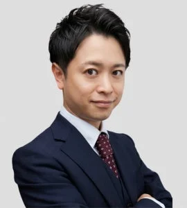 濃紺のスーツを着用し、白いシャツと柄物のネクタイを締めた日本人男性のバストアップポートレートです。カメラをまっすぐ見ており、口元にはわずかな笑みを浮かべ、プロフェッショナルな印象を与えます。