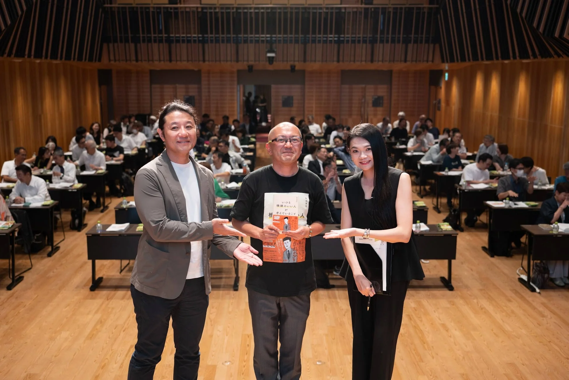 講演会かセミナーの会場で、3人の登壇者が聴衆を背景に笑顔でポーズをとっています。中央の男性は「いつも機嫌のいい人が考えていること」という書籍を手に持っており、和やかな雰囲気が伝わります。