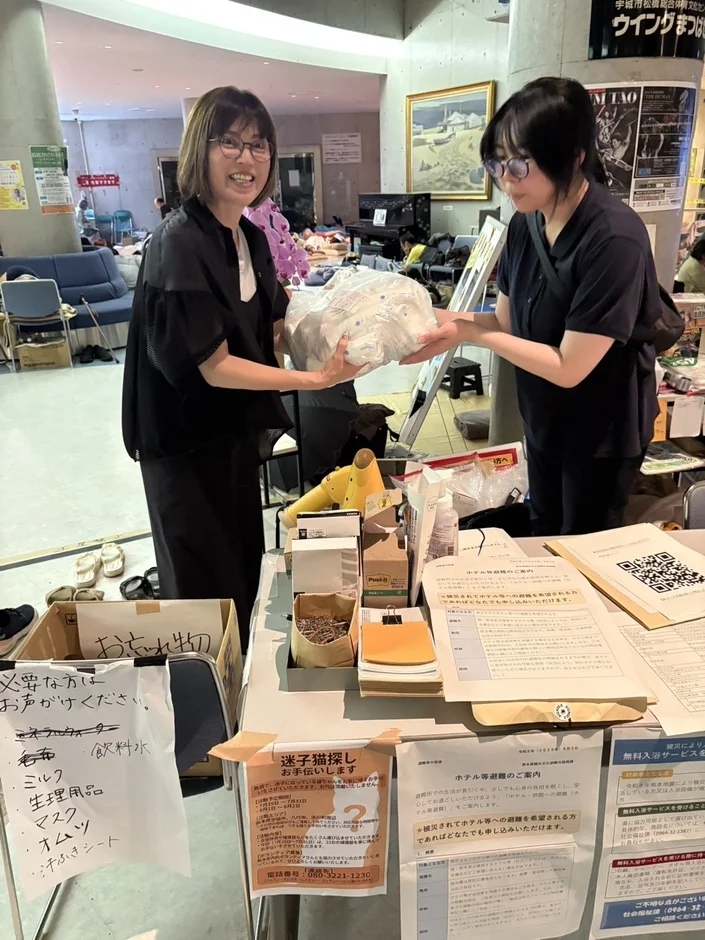 室内で女性二人が支援物資を交換。テーブルには水やマスクなどの物資リスト、迷子猫探し、ホテル避難、無料入浴サービスといった被災者支援の案内があり、多角的なサポートが行われている様子。
