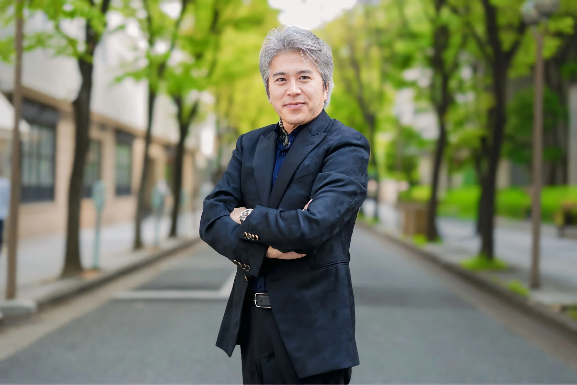 街路樹が並ぶ屋外の道で、紺色のスーツを着用し腕を組んで立つ、白髪の日本人男性のポートレート