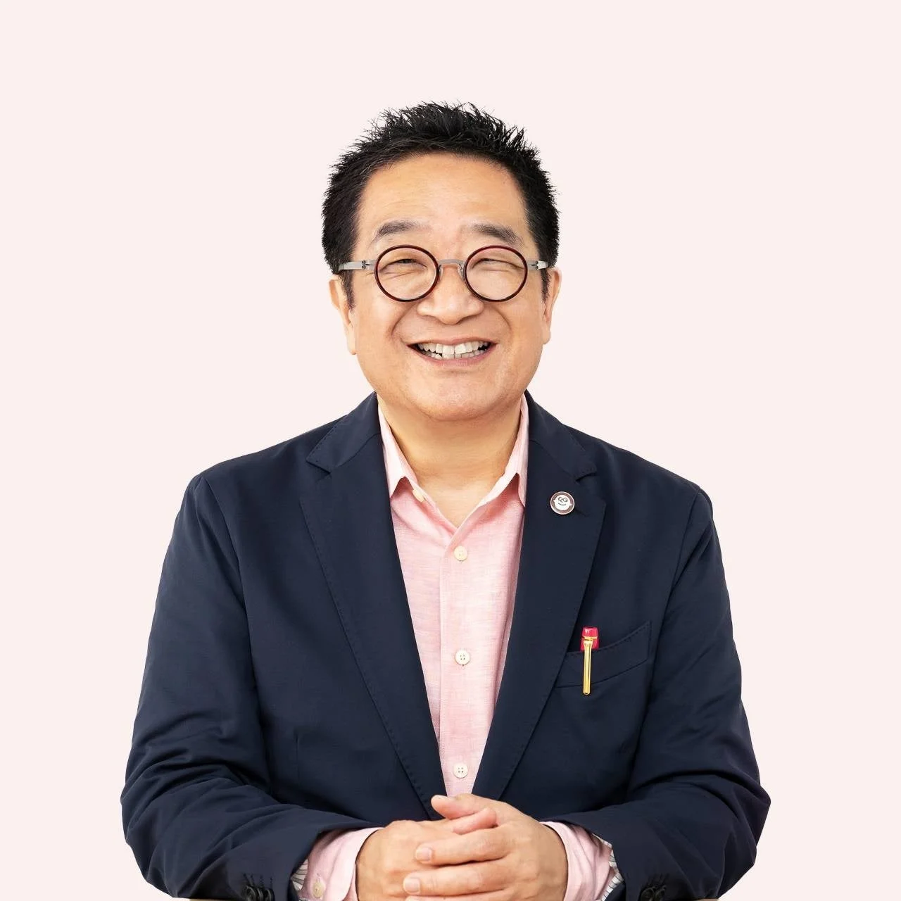 丸眼鏡をかけたアジア系の男性が、紺色のジャケットとピンクのシャツを着て、明るく微笑んでいる上半身のポートレートです。背景はシンプルな薄いピンク色です。