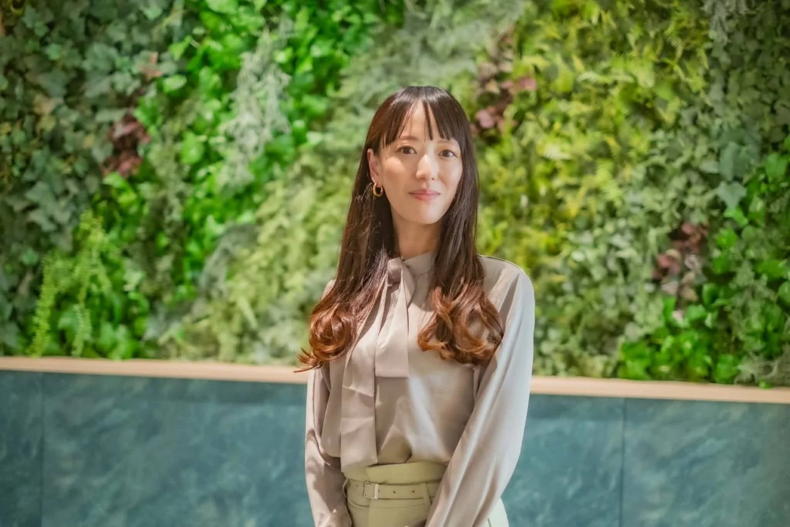 緑豊かな植物の壁を背景に、長めのウェーブヘアの若い女性が立っています。ベージュのブラウスと薄緑のパンツを着用し、穏やかな笑顔でカメラを見つめているポートレートです。