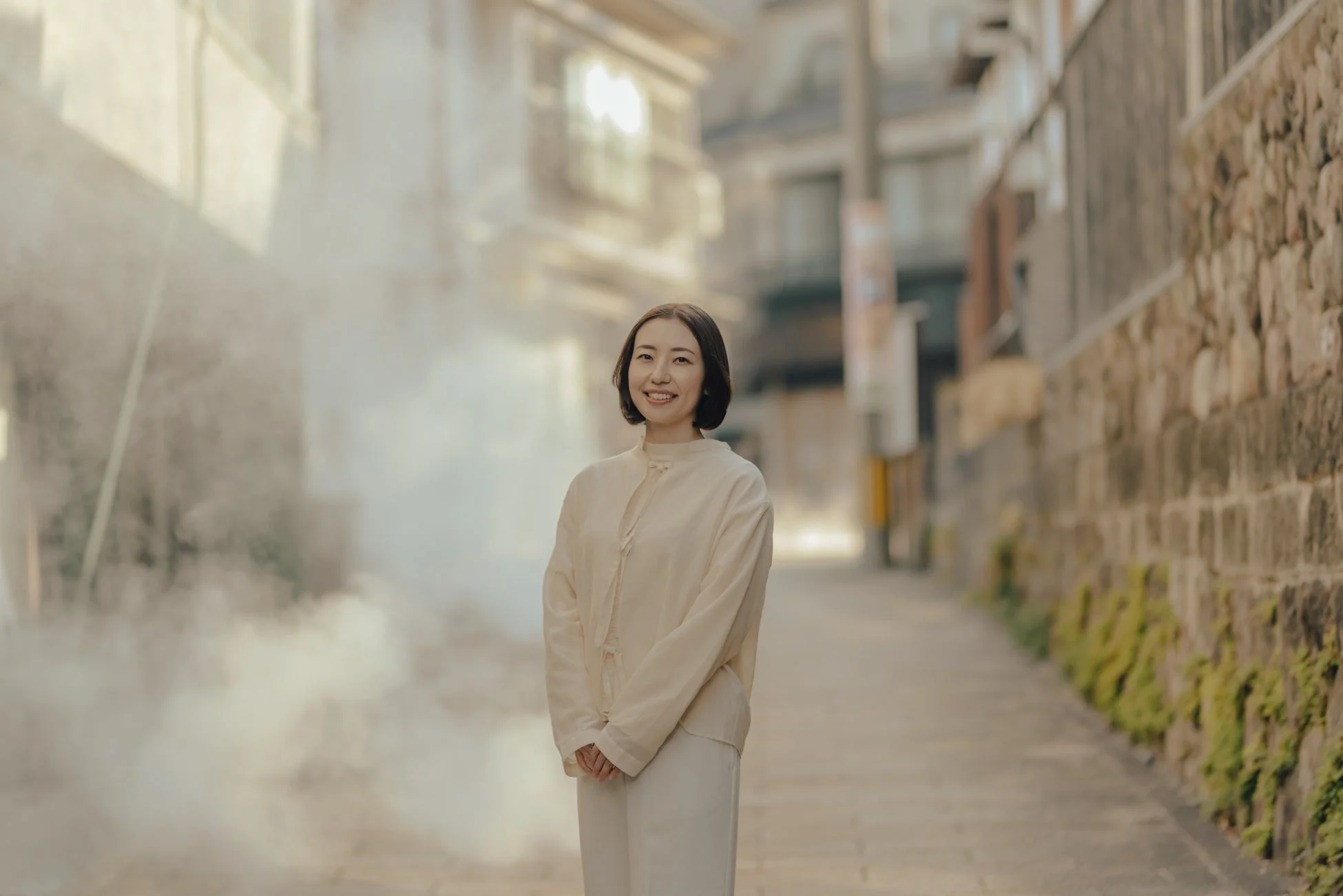 湯気が立ち込める日本の路地で、白い和装風の服を着た女性が笑顔で立っています。背景には古い建物や苔むした石垣が見え、穏やかな雰囲気が漂っています。