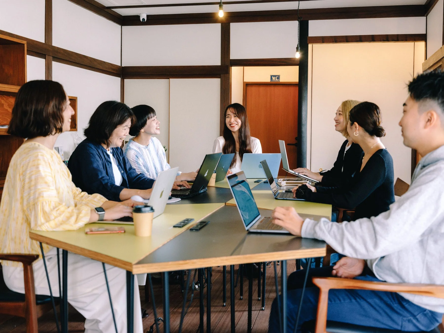 和モダンな空間で、数人の人々が大きなテーブルを囲み、ノートパソコンを使って作業している様子です。皆が笑顔で、活発な会議や共同作業が行われているように見えます。