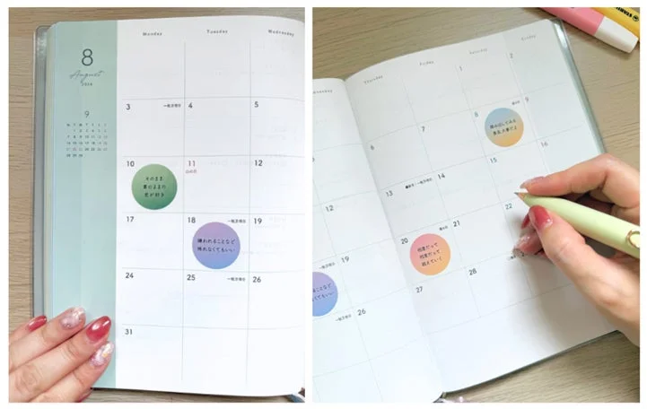 8月の手帳が開かれており、カラフルな丸いシールに自己啓発的なメッセージが書かれています。女性の指がペンを持ち、日付を指している様子が見え、計画や目標設定を行っている日常の一コマを捉えています。