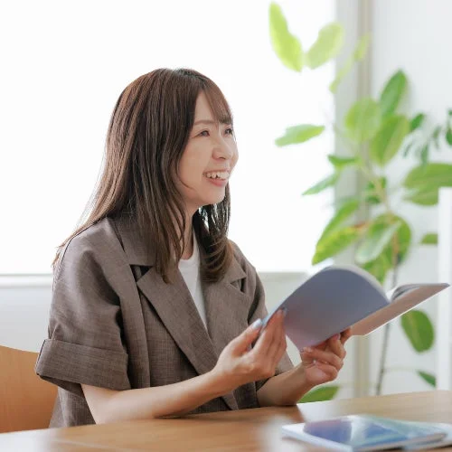 笑顔の女性が屋内のテーブルで本を開き、何かに耳を傾けているかのように横を見ています。リラックスした雰囲気で、学習やビジネスのシーンを思わせる一枚です。