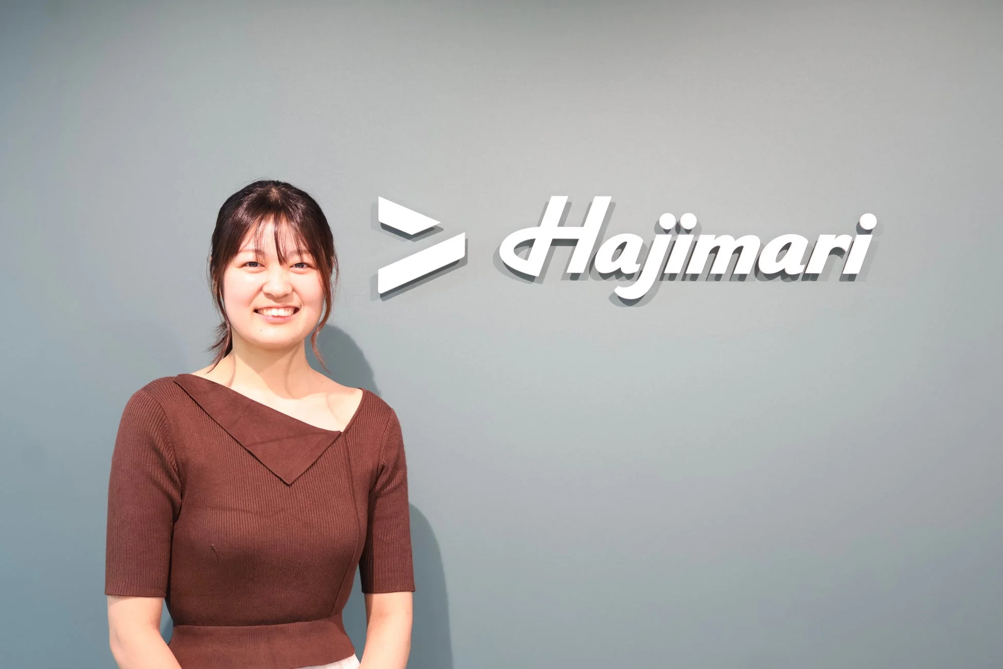 茶色のトップスを着た笑顔の女性が、灰色の壁にHajimariのロゴが掲げられた場所で立っている。