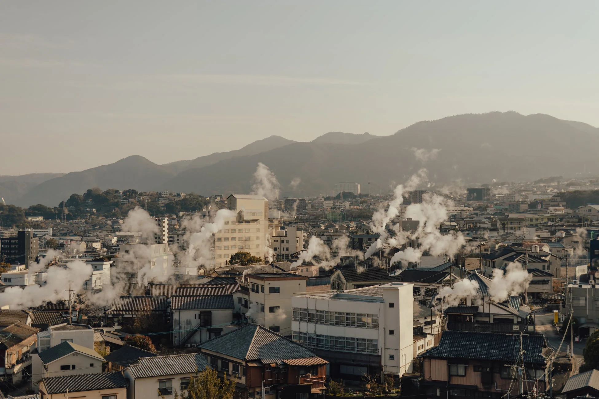 多くの建物から白い湯気が立ち上る日本の温泉街の風景。背景には山々が連なり、穏やかな日差しの中で街全体が霞がかったように見えます。